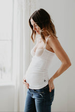 Pregnant woman wearing a white nursing tank top and blue jeans standing in a bright room with a window.